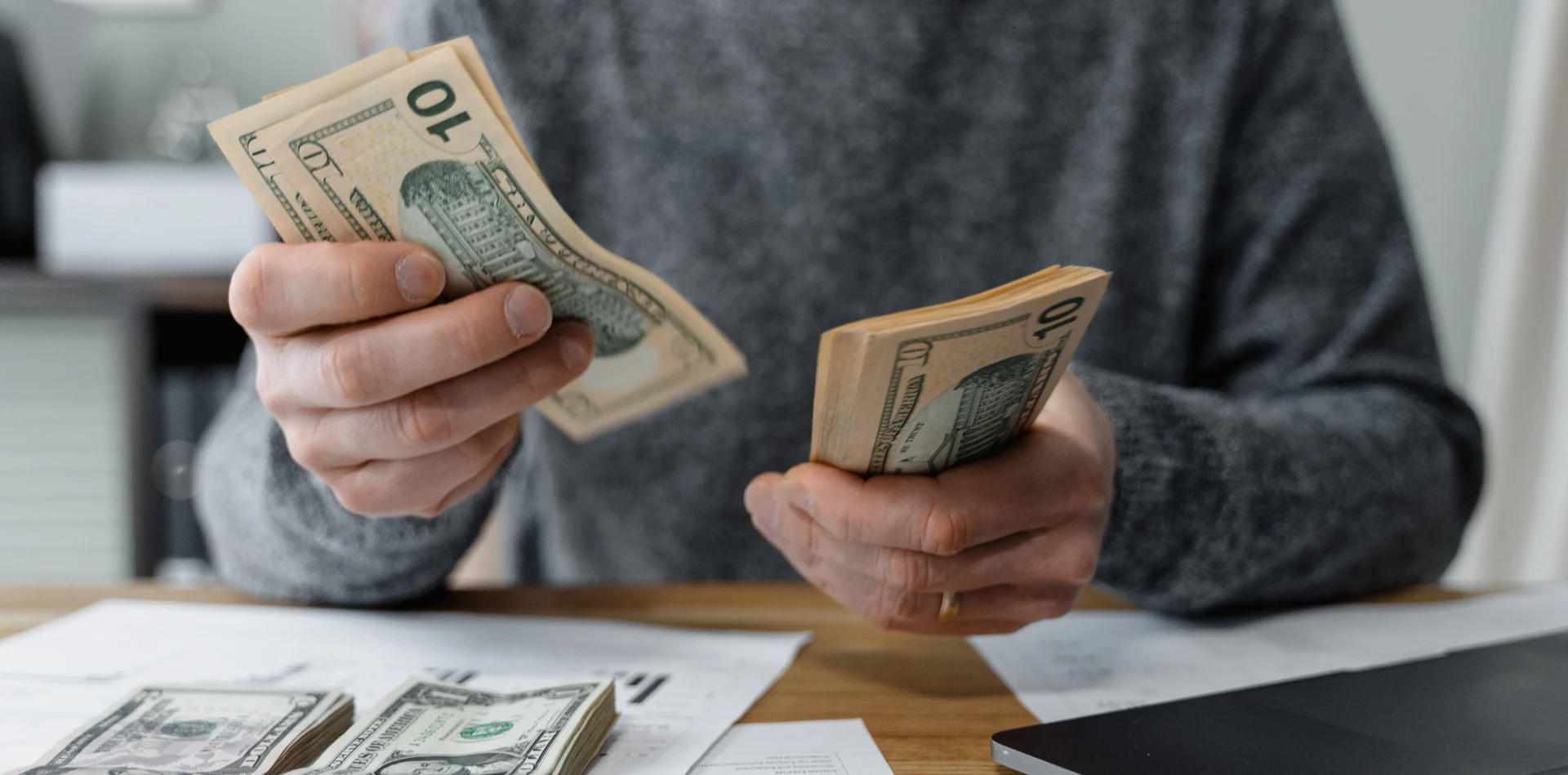 Person counting US ten-dollar bills at a desk with papers and a laptop.