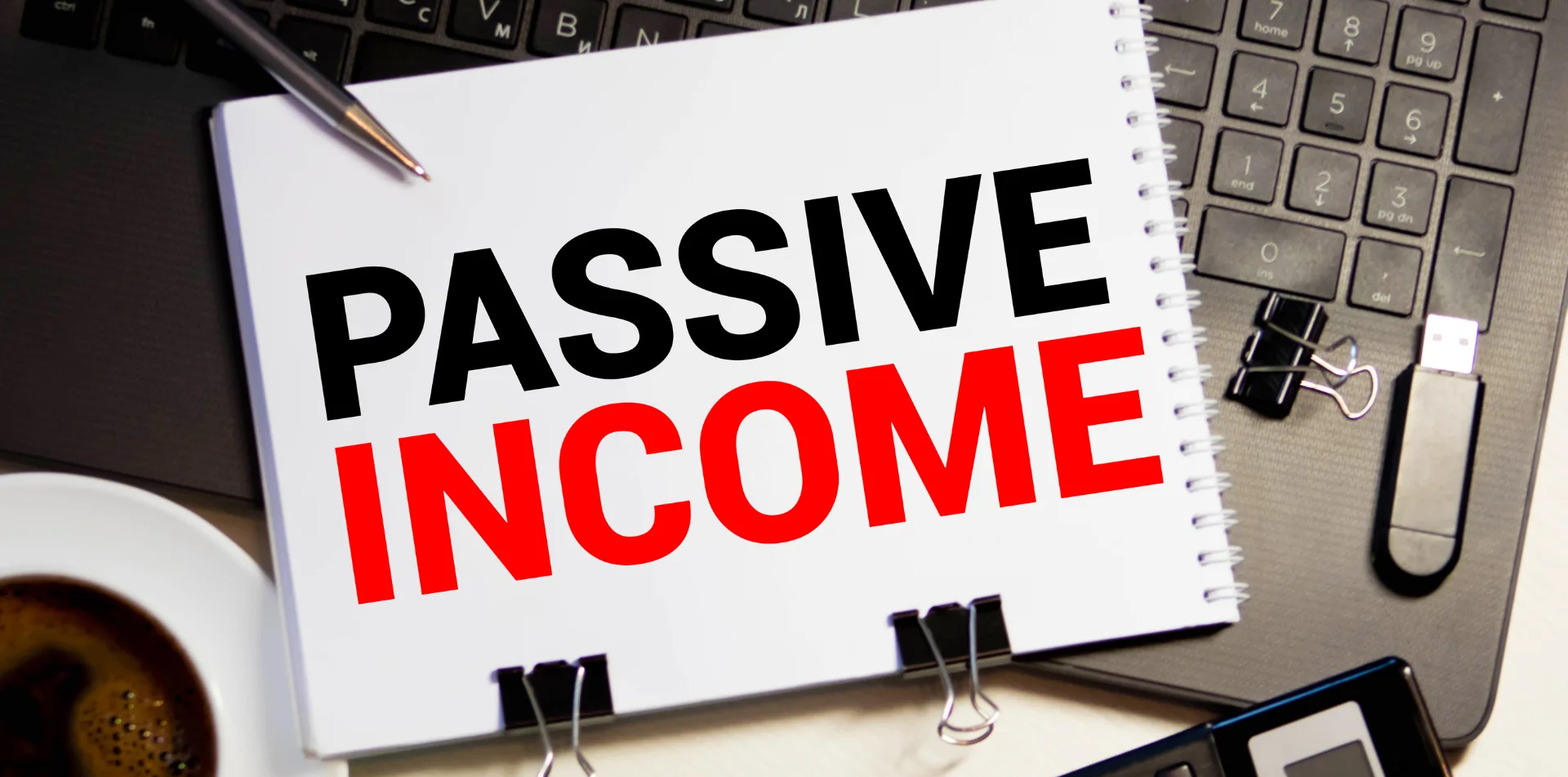 Notebook with "PASSIVE INCOME" text, pen, and coffee cup on a keyboard.